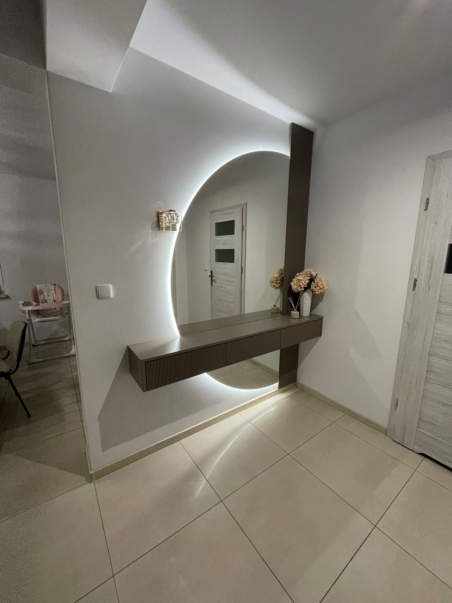 Nowoczesna aranżacja przedpokoju z okrągłym lustrem podświetlanym LED, konsolą z szufladami i dekoracyjnym oświetleniem ściennym. W tle widoczny pokój z krzesełkiem do karmienia.