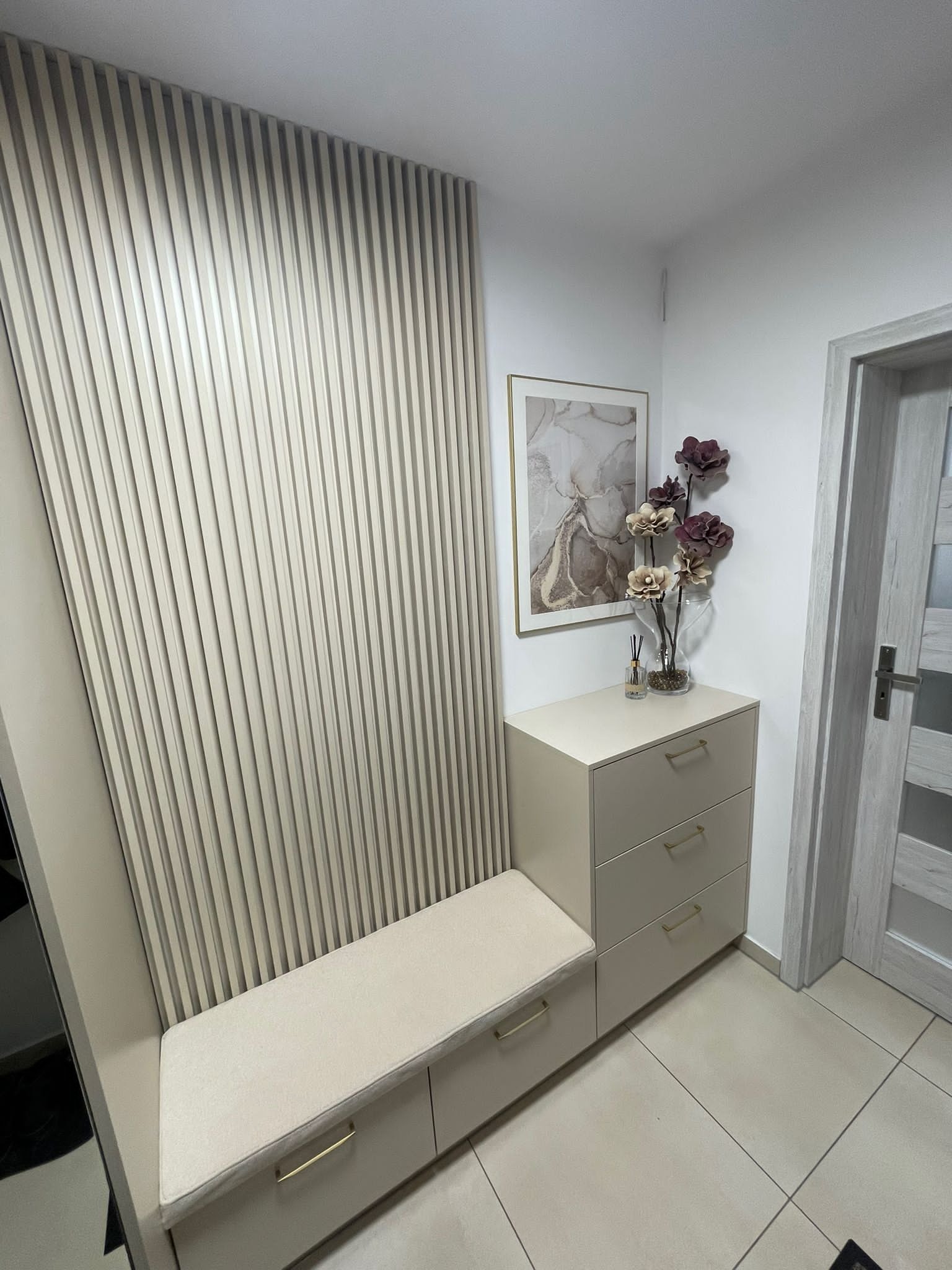 Beżowa zabudowa przedpokoju z siedziskiem, szufladami i panelem ściennym z pionowych listew, dekoracje w postaci obrazu i kwiatów.