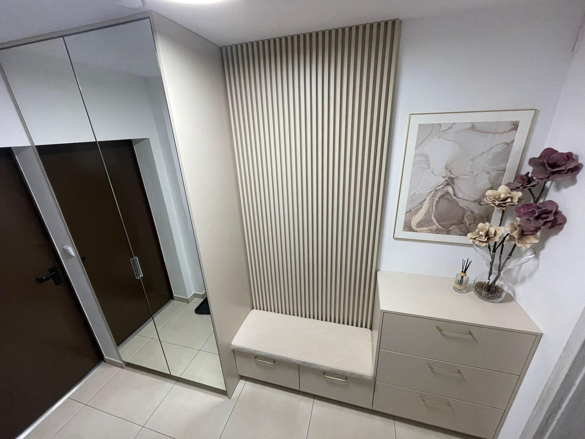 Wnętrze przedpokoju z szafą z lustrem, panelem ściennym z pionowych listew, komodą i siedziskiem, dekorowane obrazem w ramie i kwiatami.