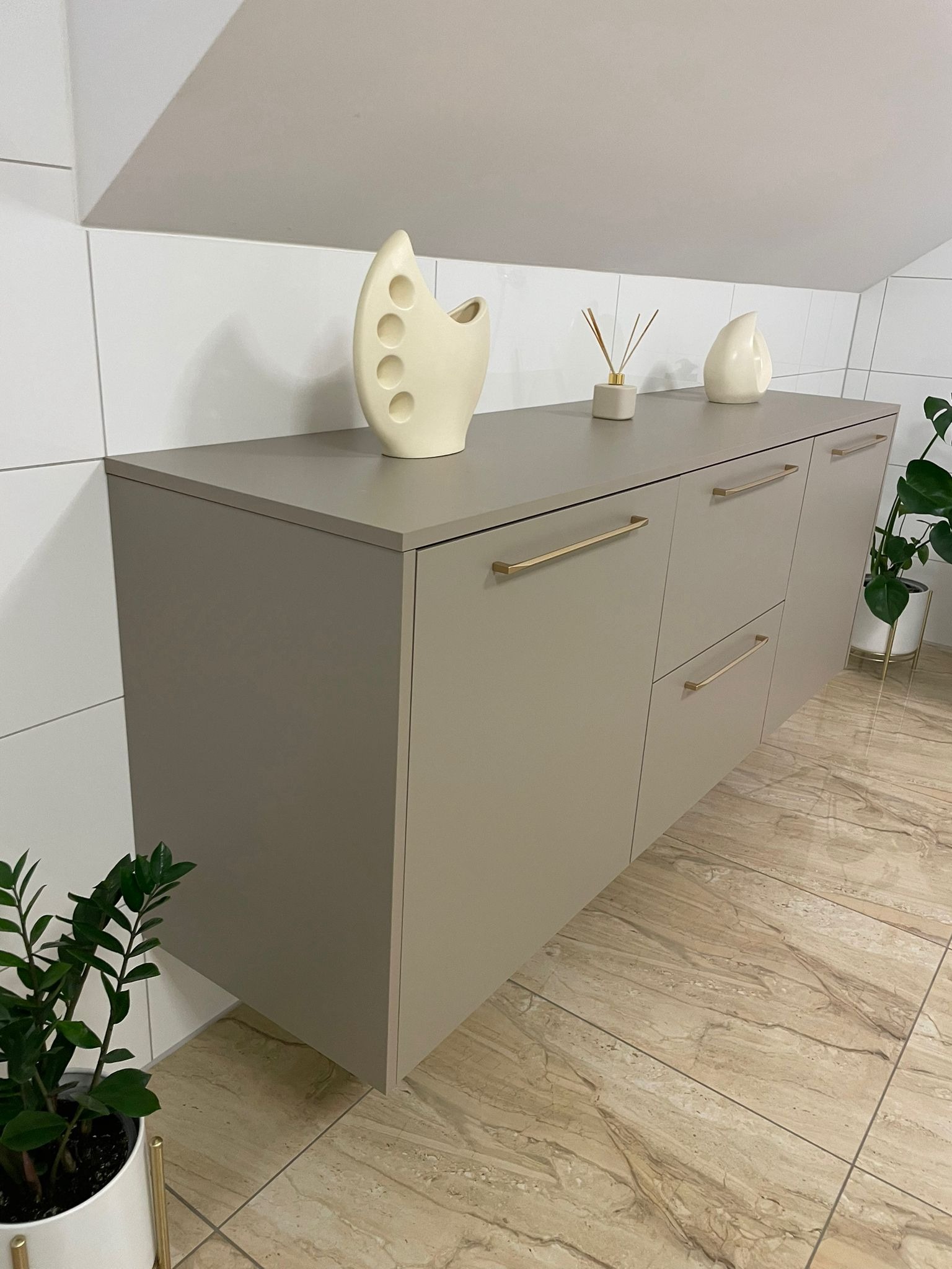 Beżowa szafka RTV z długimi złotymi uchwytami, na blacie dekoracyjne wazony i zapach do wnętrz, na tle białych płytek i marmurowej podłogi.