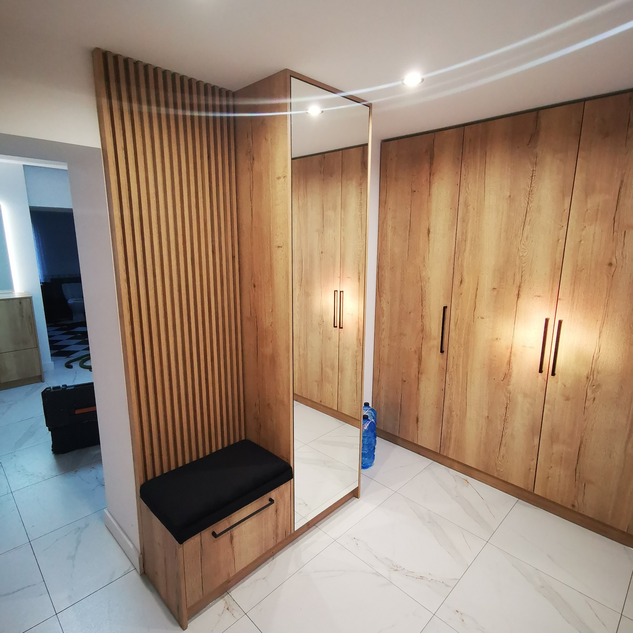 Nowoczesna zabudowa przedpokoju z drewnianymi lamelami, siedziskiem z czarną tapicerką i lustrem, obok duża szafa z drewnopodobnymi frontami.