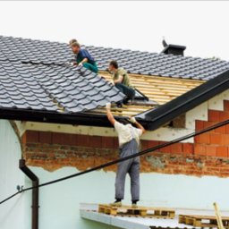 Dach serwis - Trzech mężczyzn pracuje na dachu domu. Dwóch układa ciemnoszarą dachówkę, a trzeci stoi na podeście z palet i podaje materiały. Widoczne elementy więźby i fragment ściany z cegły.