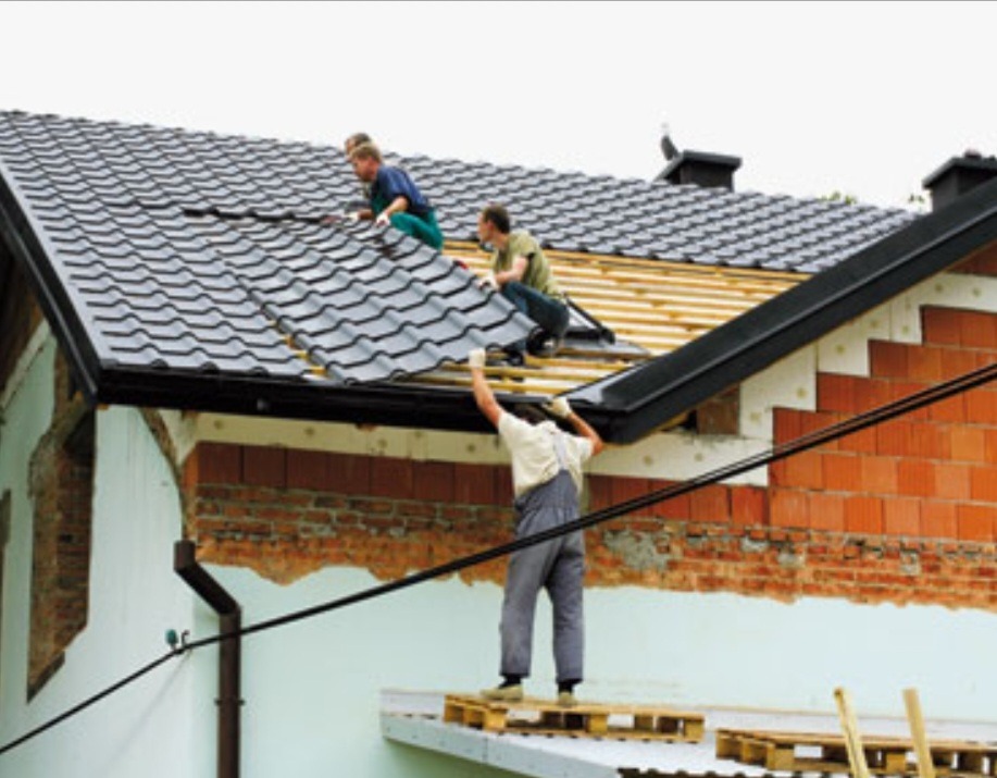 Trzech mężczyzn pracuje na dachu domu. Dwóch układa ciemnoszarą dachówkę, a trzeci stoi na podeście z palet i podaje materiały. Widoczne elementy więźby i fragment ściany z cegły.