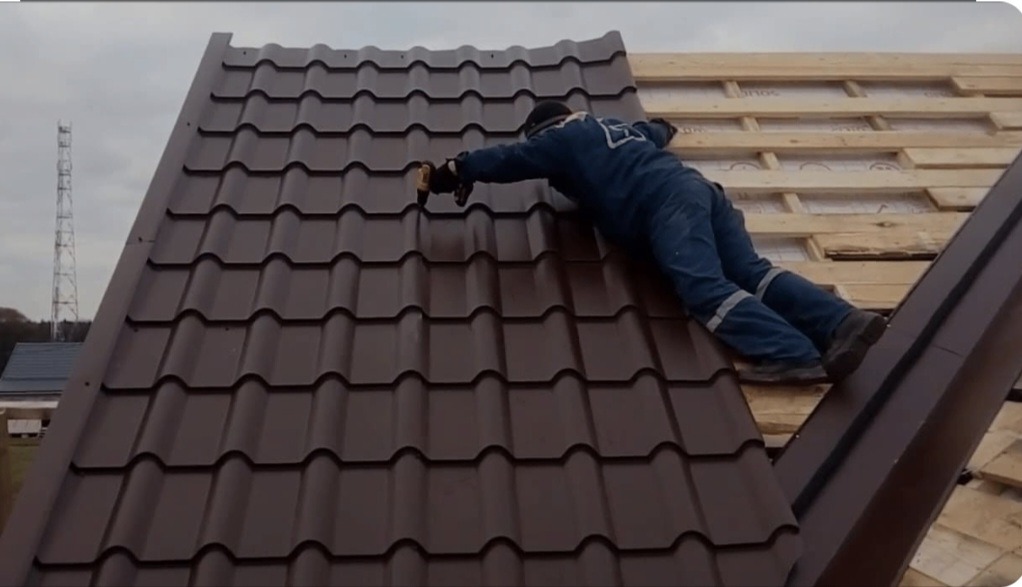 Pracownik w niebieskim kombinezonie mocuje brązowe panele dachowe do drewnianej konstrukcji dachu za pomocą wkrętarki.