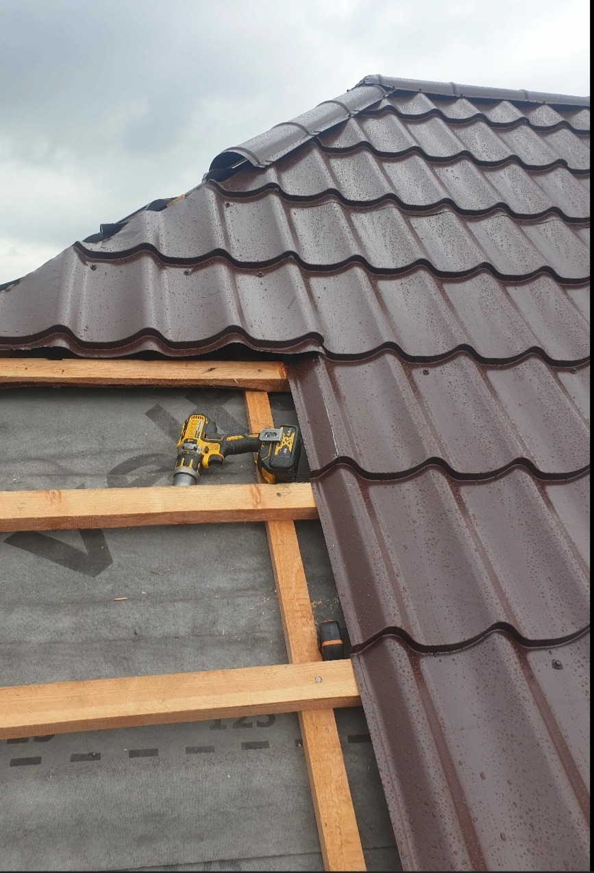 Montaż brązowej blachodachówki na drewnianej konstrukcji pod deszczowym niebem, wiertarko-wkrętarka DeWalt na tle czarnej membrany dachowej.