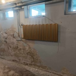 Bemal Bartłomiej Sikora - Ściana w trakcie remontu z widocznymi uszkodzeniami i częściowo nałożoną gładzią, z zamontowanym panelem dekoracyjnym. Widoczne okna i instalacje.