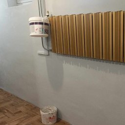 Bemal Bartłomiej Sikora - Świeżo pomalowane na szaro wnętrze z drewnianą dekoracją ścienną, wiaderkiem farby i metalową szafą. Podłoga wyłożona panelami imitującymi drewno.