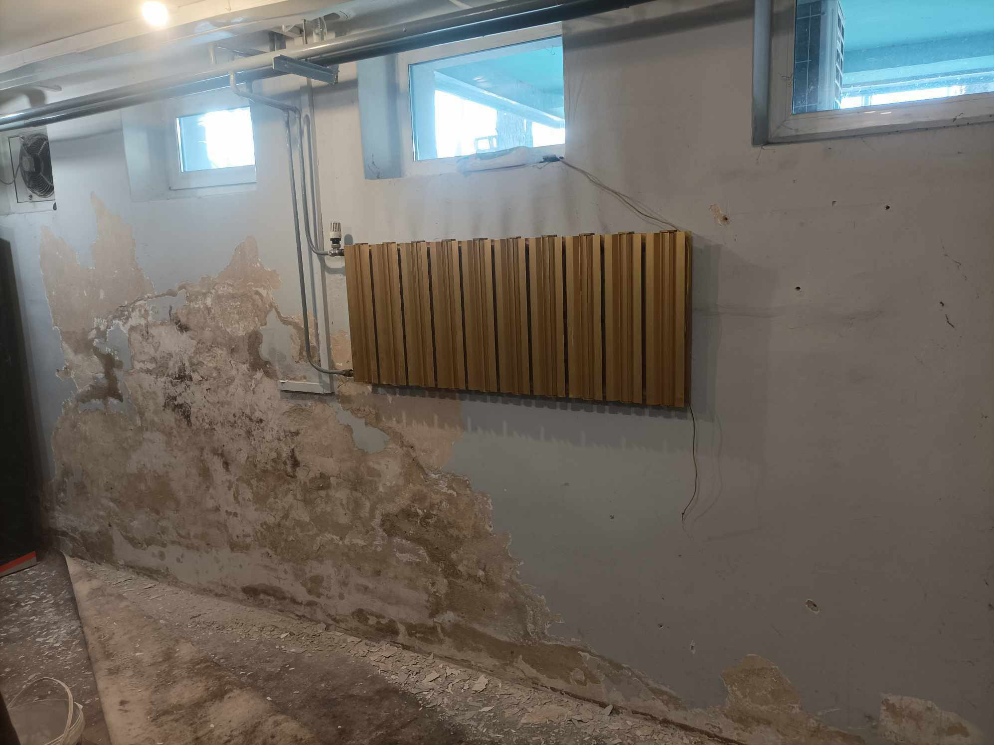 Ściana w trakcie remontu z widocznymi uszkodzeniami i częściowo nałożoną gładzią, z zamontowanym panelem dekoracyjnym. Widoczne okna i instalacje.