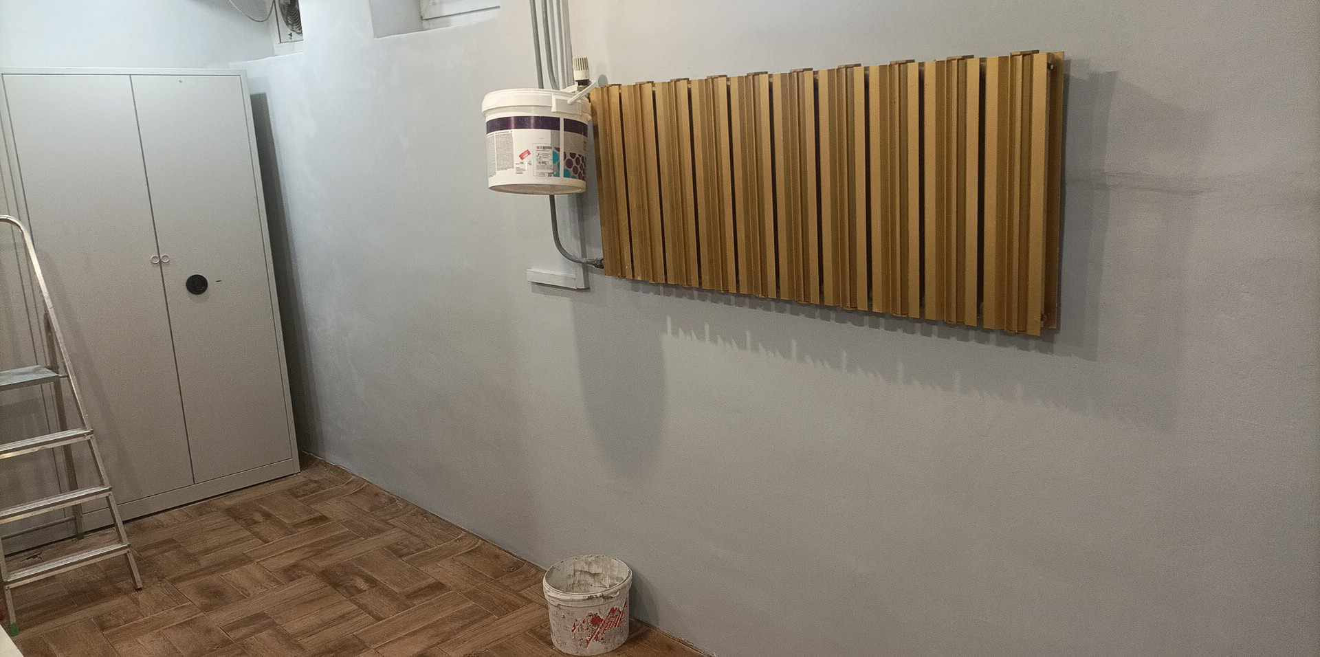 Świeżo pomalowane na szaro wnętrze z drewnianą dekoracją ścienną, wiaderkiem farby i metalową szafą. Podłoga wyłożona panelami imitującymi drewno.