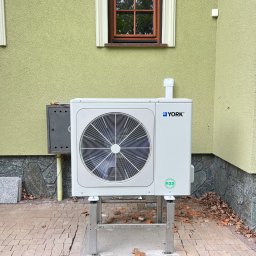 T- service Oleh Shablovskyi - Biała pompa ciepła York R32, zamontowana na metalowym stelażu przy budynku z zieloną elewacją i brązowym oknem. Widoczne kostki brukowe wokół.