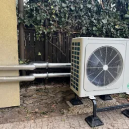 Biała pompa ciepła York zamontowana na zewnątrz budynku z izolowanymi rurami, na tle ogrodzenia porośniętego bluszczem i kostki brukowej.
