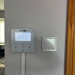 T- service Oleh Shablovskyi - Panel sterowania ogrzewaniem marki York zamontowany na ścianie obok włącznika światła i drewnianej framugi drzwi. Widoczny ekran z ustawieniami i przyciskami.