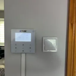 Panel sterowania ogrzewaniem marki York zamontowany na ścianie obok włącznika światła i drewnianej framugi drzwi. Widoczny ekran z ustawieniami i przyciskami.