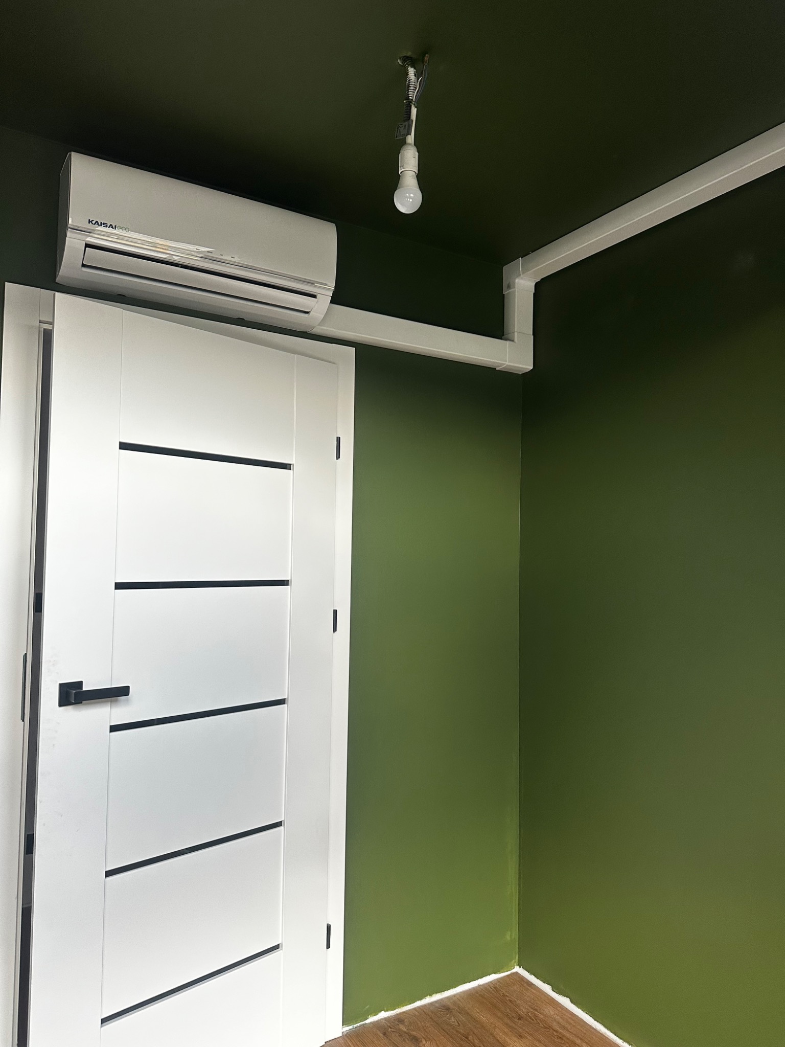 Wnętrze z białą klimatyzacją Kaisai Eco nad drzwiami, biała instalacja elektryczna pod sufitem, zielone ściany i drewniana podłoga. Minimalistyczny design.