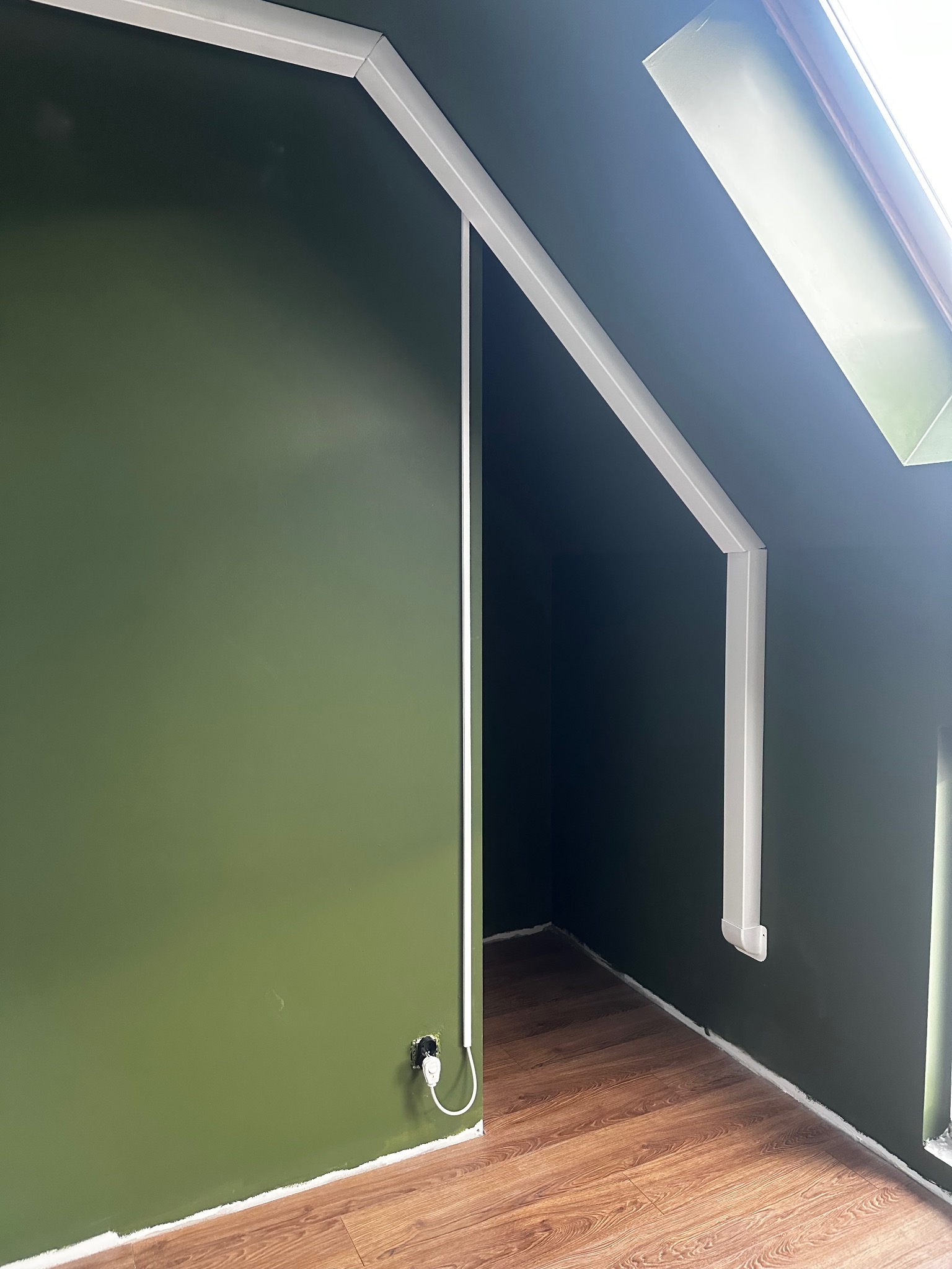 Zielona ściana na poddaszu z listwami przypodłogowymi i kablem elektrycznym przy gniazdku. Widoczny fragment okna dachowego i podłoga z paneli.