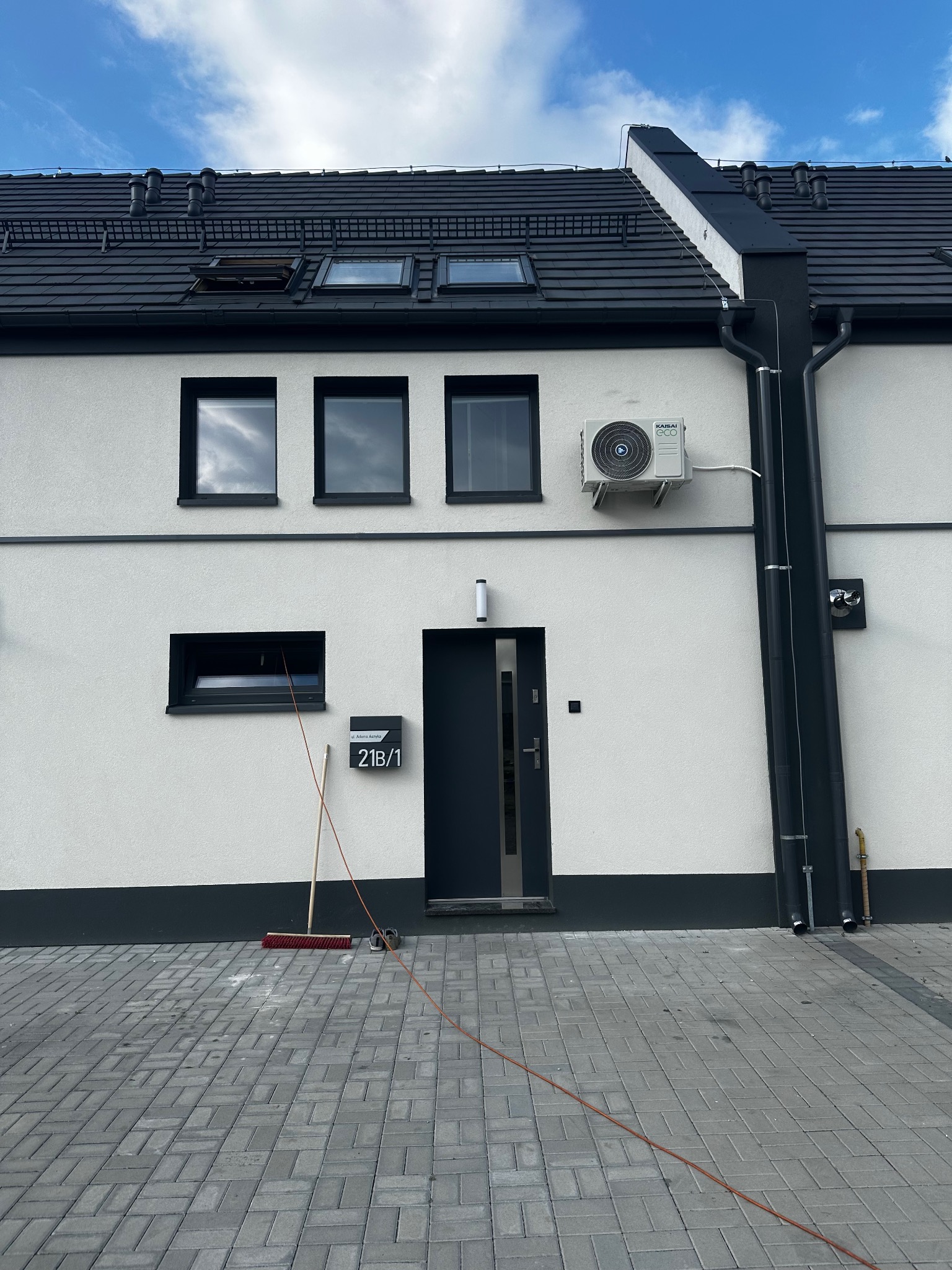 Biały dom z szarą stolarką i dachem, widoczna boczna ściana z klimatyzatorem Kaisai Eco, rynny, numer domu 21B/1 i kostka brukowa. Dom po instalacji.