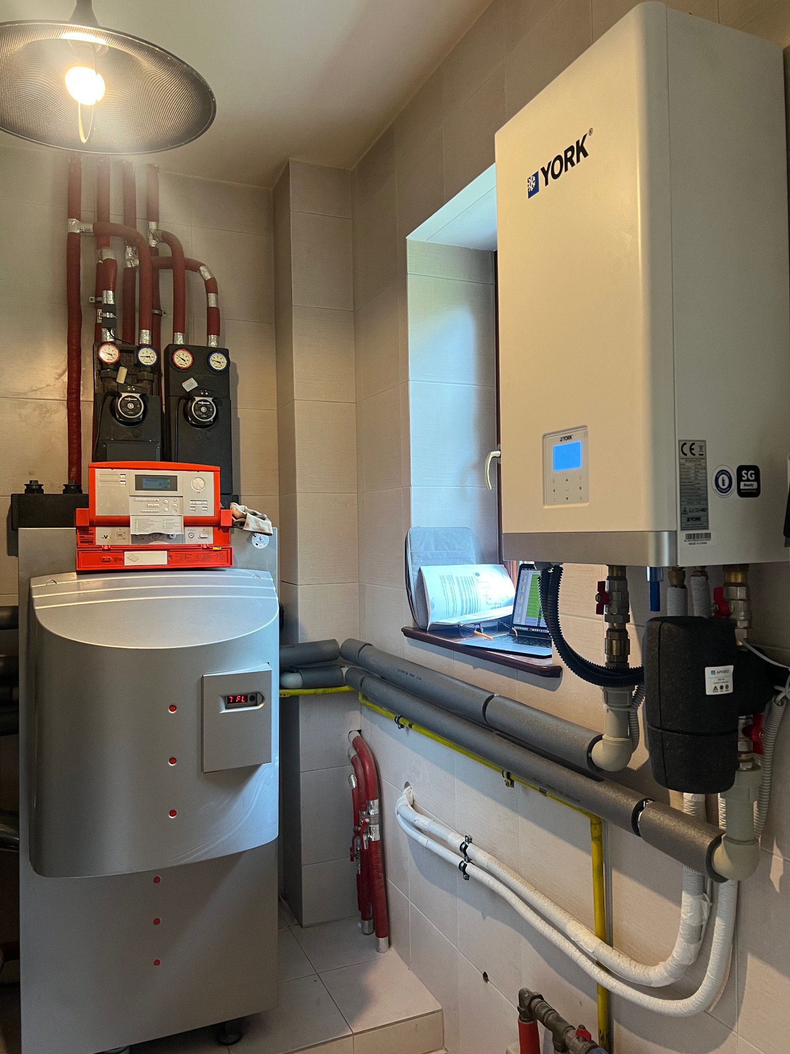 Nowoczesna kotłownia z piecem gazowym York, widoczne rury instalacji grzewczej, manometry i sterownik. Na parapecie laptop i dokumentacja techniczna.