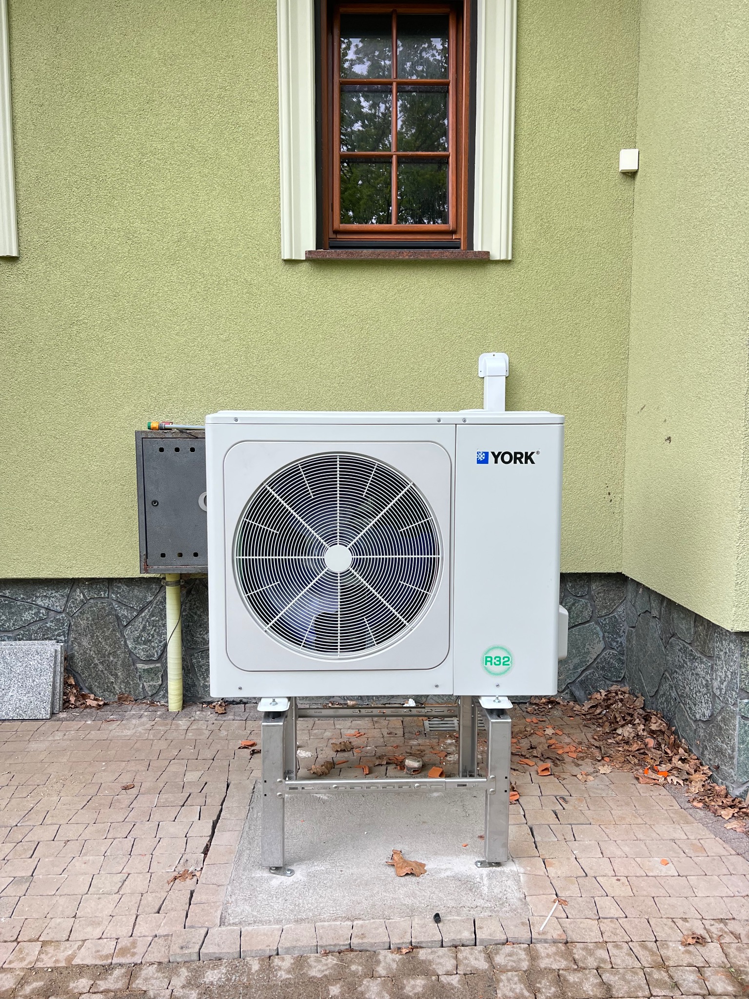 Biała pompa ciepła York R32, zamontowana na metalowym stelażu przy budynku z zieloną elewacją i brązowym oknem. Widoczne kostki brukowe wokół.