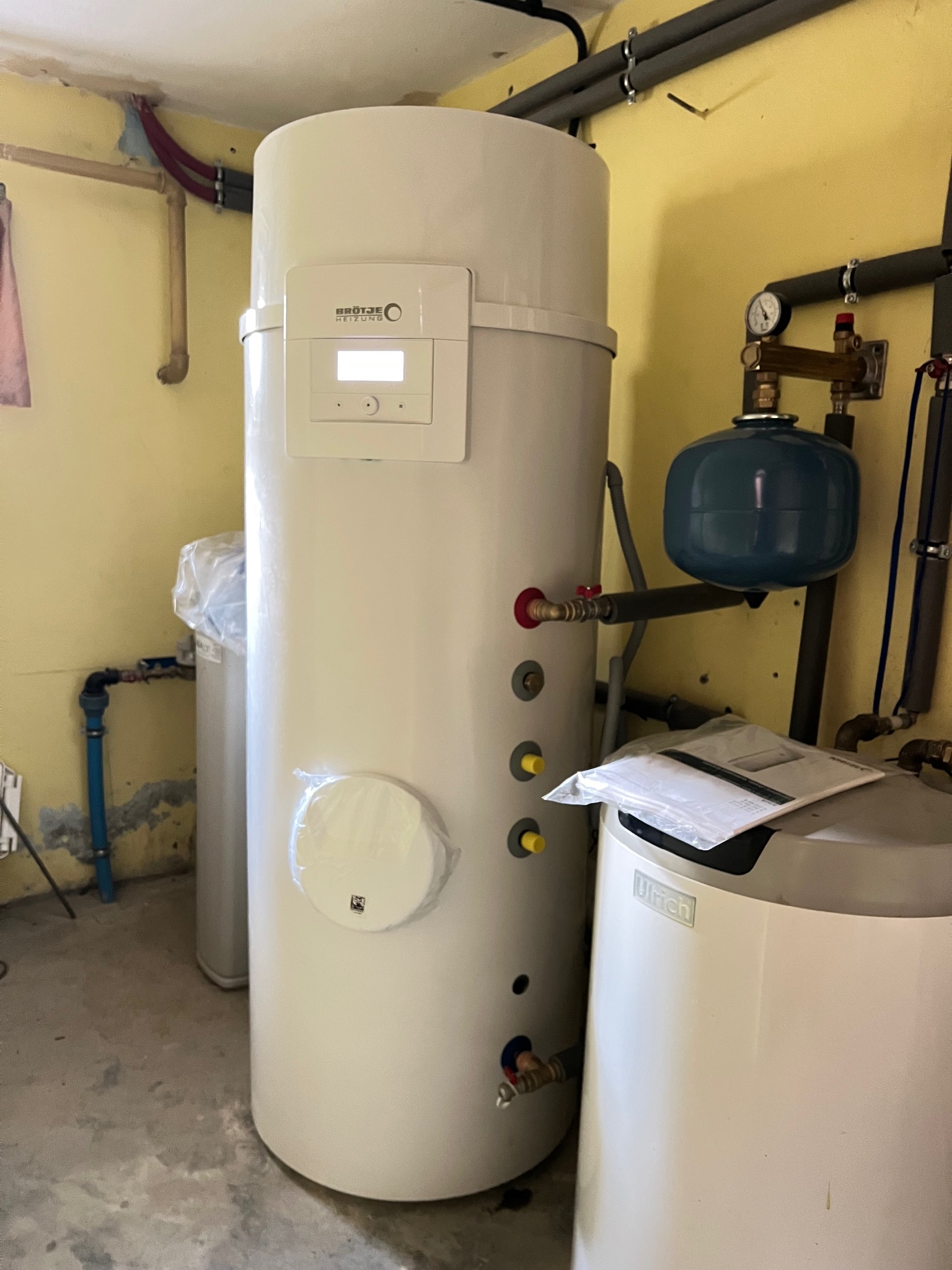 Biały, cylindryczny zbiornik Brötje z panelem sterowania, obok mniejszy zbiornik Ulrich, instalacja hydrauliczna na żółtej ścianie. Elementy instalacji grzewczej w kotłowni.