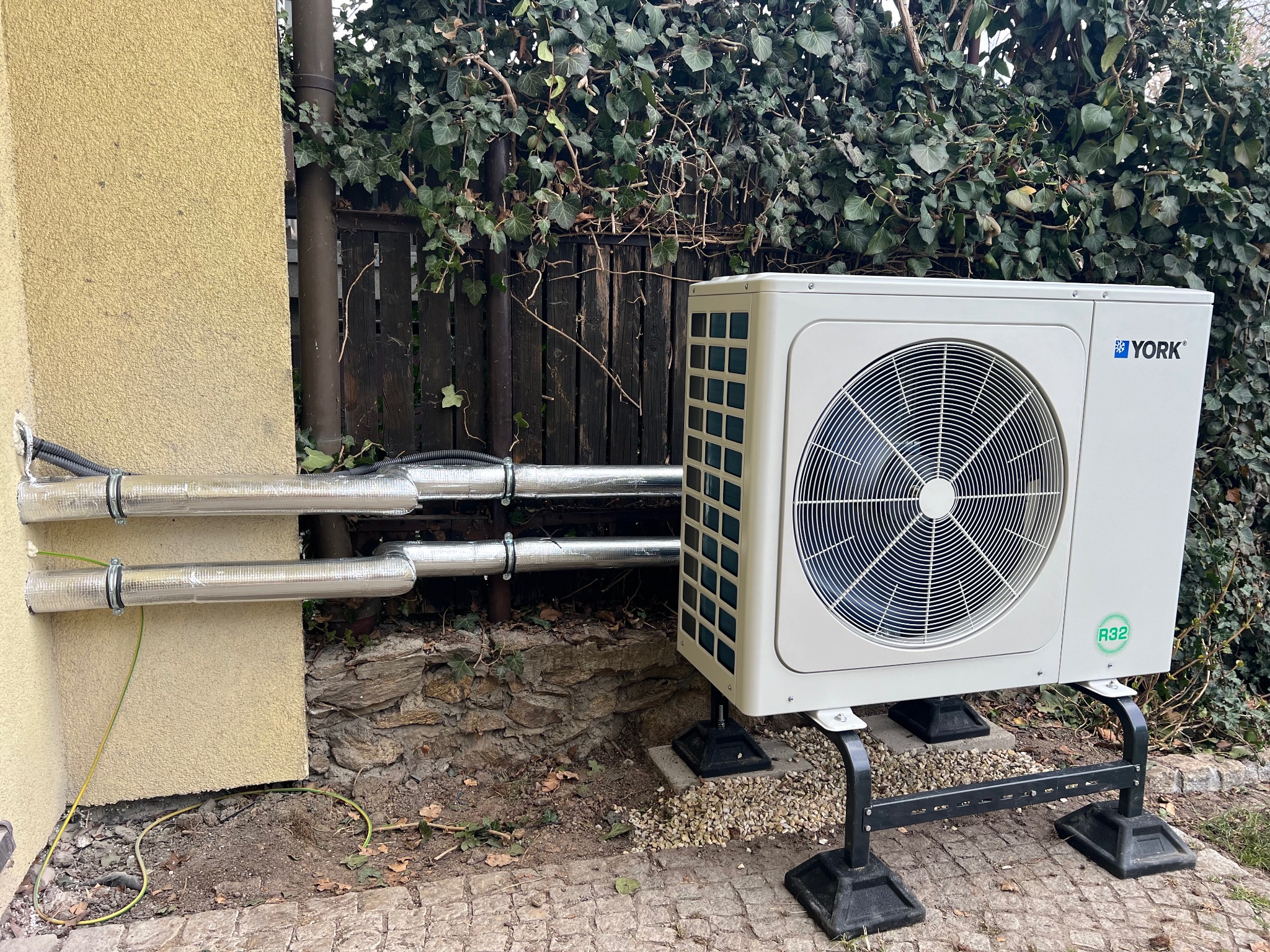 Biała pompa ciepła York zamontowana na zewnątrz budynku z izolowanymi rurami, na tle ogrodzenia porośniętego bluszczem i kostki brukowej.
