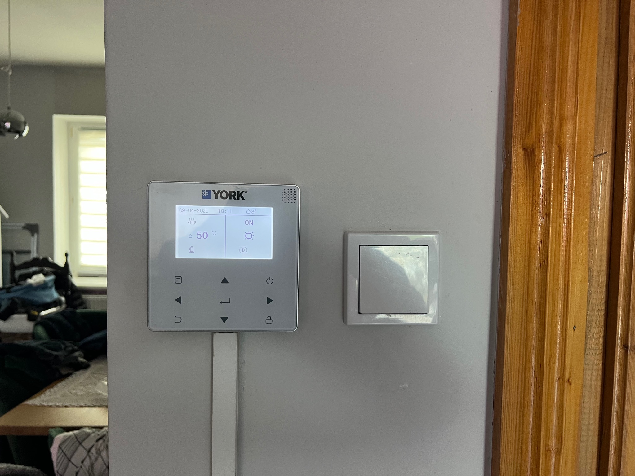 Panel sterowania ogrzewaniem marki York zamontowany na ścianie obok włącznika światła i drewnianej framugi drzwi. Widoczny ekran z ustawieniami i przyciskami.