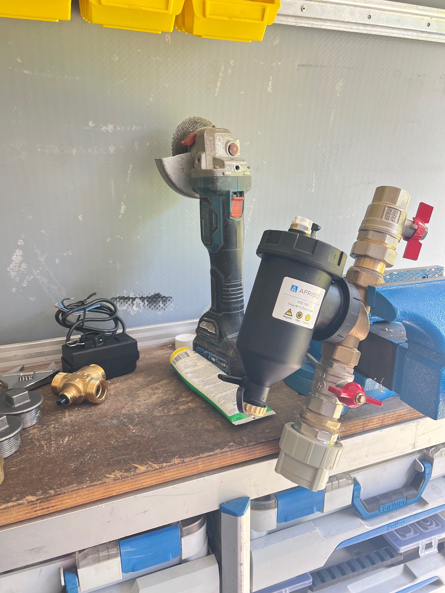 Filtr magnetyczny AFRISO ADS 160 zamontowany na rurach w imadle, obok szlifierka kątowa i narzędzia na warsztatowym stole. Widoczne złącza i zawory.