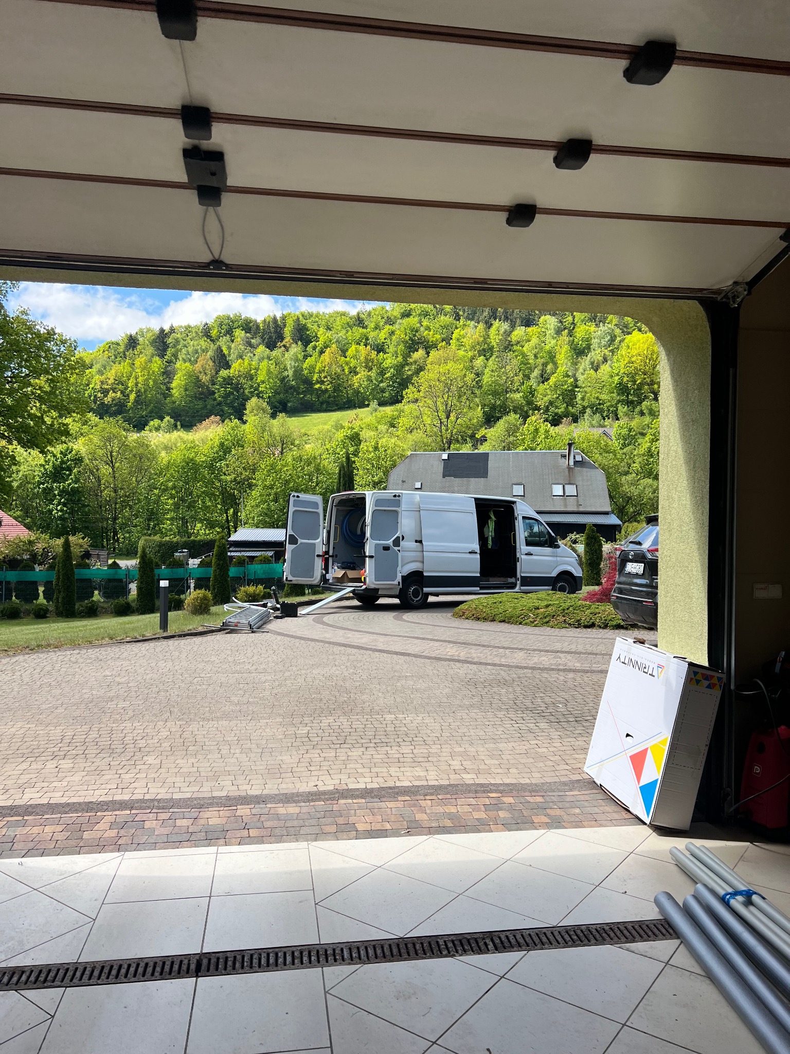 Widok z garażu na pojazd instalacyjny z otwartymi drzwiami, sprzęt i pudełko Trinity na podjeździe, w tle zieleń. Świeże powietrze i nowoczesny sprzęt.