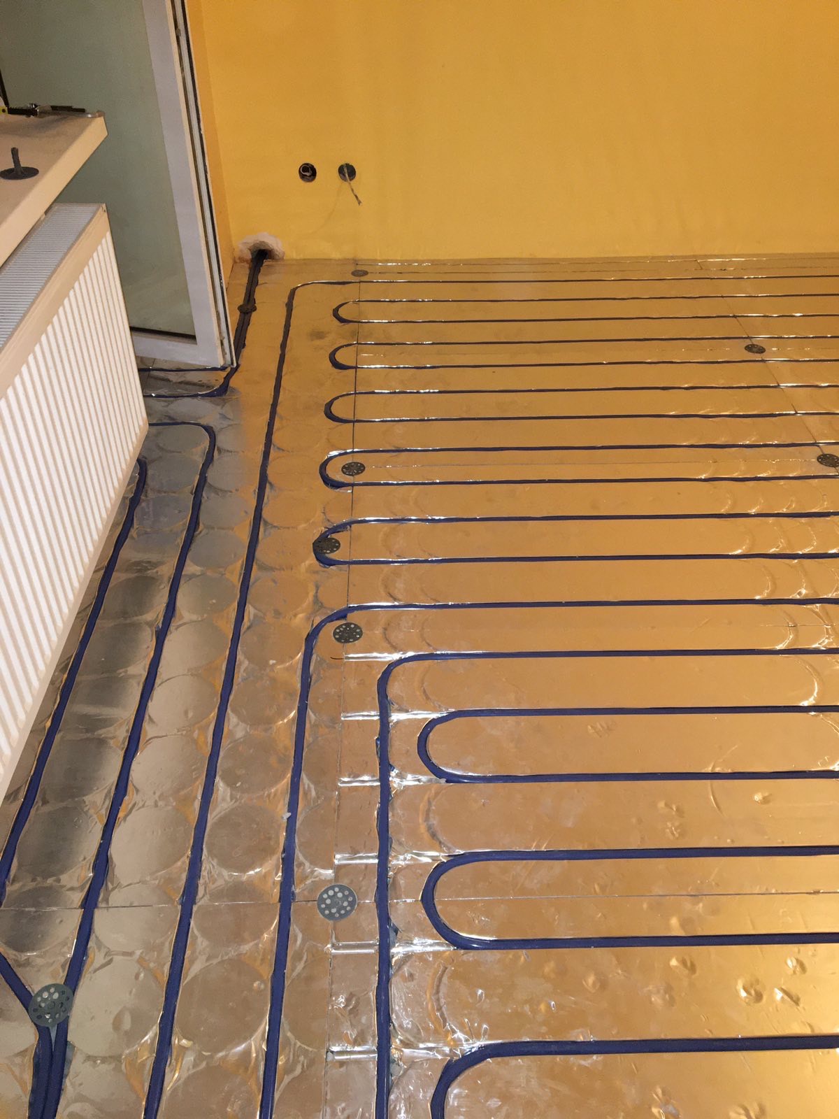 Błękitne rurki ogrzewania podłogowego ułożone na srebrnej folii izolacyjnej, widoczne uchwyty mocujące, fragment grzejnika. Żółta ściana z gniazdkami w tle.