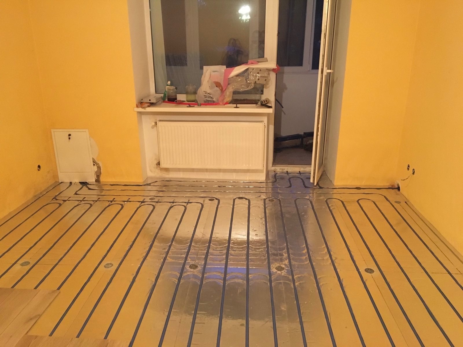 Montaż ogrzewania podłogowego z folią aluminiową w pokoju o żółtych ścianach, widoczny grzejnik pod oknem i instalacja elektryczna w ścianie.