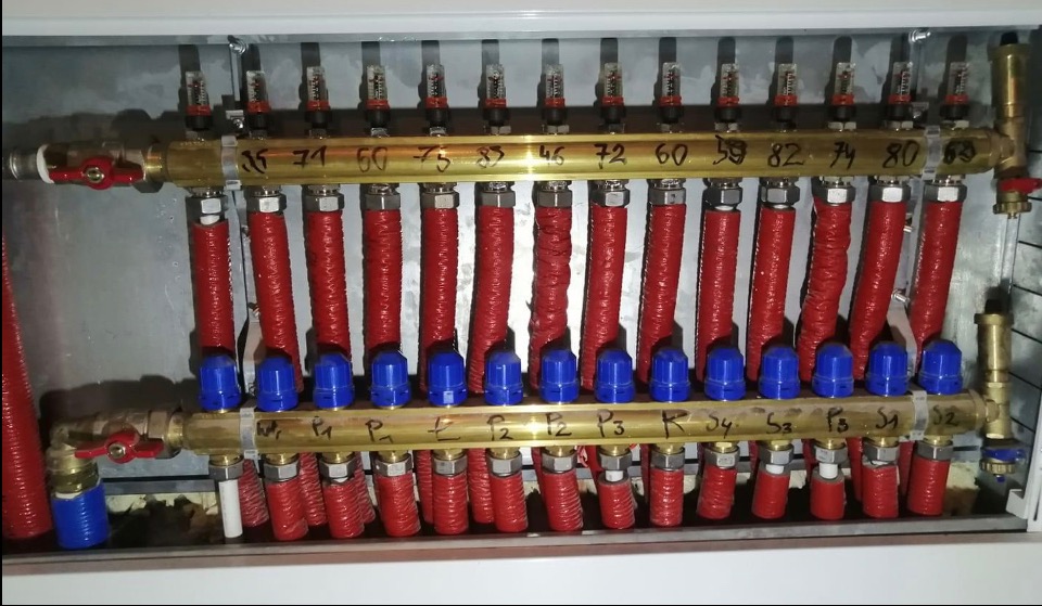 Rozdzielacz ogrzewania podłogowego z rurami w izolacji, zaworami z niebieskimi pokrętłami i oznaczonymi sekcjami. Widoczne numery i litery na elementach instalacji.