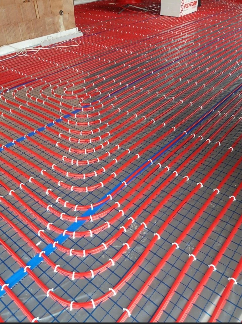 Czerwone rury ogrzewania podłogowego ułożone na izolacji z folią w szarą kratkę, przymocowane białymi uchwytami, widoczne niebieskie paski mocujące.