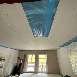 Komplexbud Yuliia Hedikova - Montaż płyt gipsowo-kartonowych na suficie podwieszanym, zabezpieczenie folią okien i ścian przed zabrudzeniem, widoczni dwaj pracownicy w trakcie pracy.