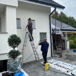 Komplexbud Yuliia Hedikova - Dwóch mężczyzn czyści białą elewację domu. Jeden stoi na drabinie z przedłużką do wałka, drugi obsługuje myjkę ciśnieniową na ziemi. Widoczne rusztowanie z boku budynku i donica z drzewkiem.