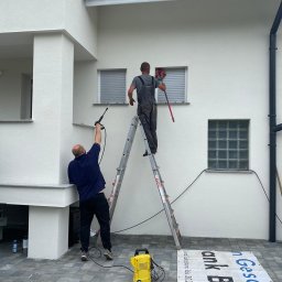 Komplexbud Yuliia Hedikova - Dwóch mężczyzn czyści elewację domu: jeden z myjką ciśnieniową z dołu, drugi z szczotką na kiju stojąc na drabinie, widoczna kostka brukowa i baner reklamowy.