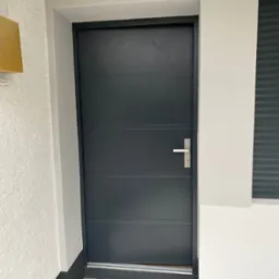 Nowoczesne, grafitowe drzwi wejściowe z poziomymi panelami i srebrną klamką, osadzone w białym murze z teksturą, fragment okna z żaluzjami w tle.