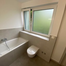 Komplexbud Yuliia Hedikova - Biała wanna, podwieszana toaleta i okno z mleczną szybą w wykończonej łazience z szarymi płytkami.