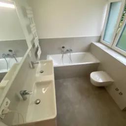 Nowoczesna łazienka z dwiema umywalkami, wanną i wiszącą toaletą, utrzymana w szaro-białej kolorystyce, z grzejnikiem drabinkowym i dużym oknem.