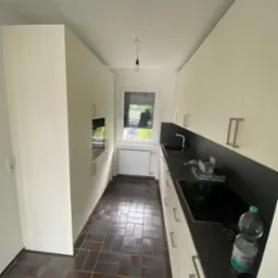 Wnętrze kuchni z brązową podłogą z płytek, białymi szafkami z długimi, srebrnymi uchwytami i czarnym blatem, widok z perspektywy wejścia.