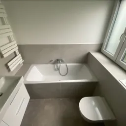 Nowoczesna łazienka w neutralnych barwach z białą armaturą: wanna z baterią, umywalka z szafką i toaleta. Na ścianie grzejnik drabinkowy.