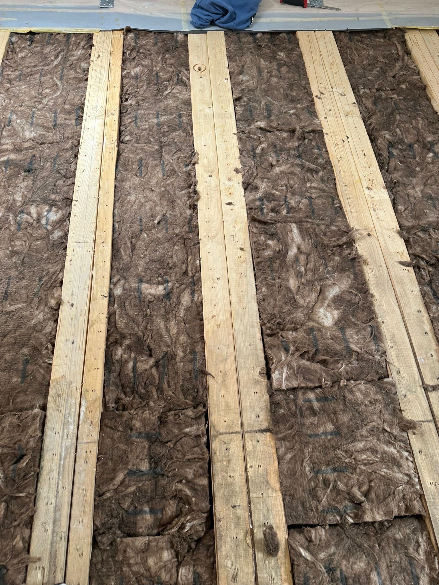 Izolacja z wełny mineralnej pomiędzy drewnianymi legarami podłogowymi, widoczne ślady po cięciu wełny, na górze fragment niebieskiej odzieży roboczej.