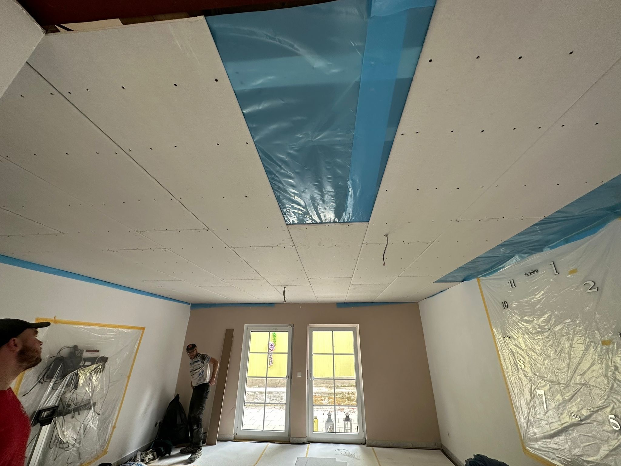 Montaż płyt gipsowo-kartonowych na suficie podwieszanym, zabezpieczenie folią okien i ścian przed zabrudzeniem, widoczni dwaj pracownicy w trakcie pracy.