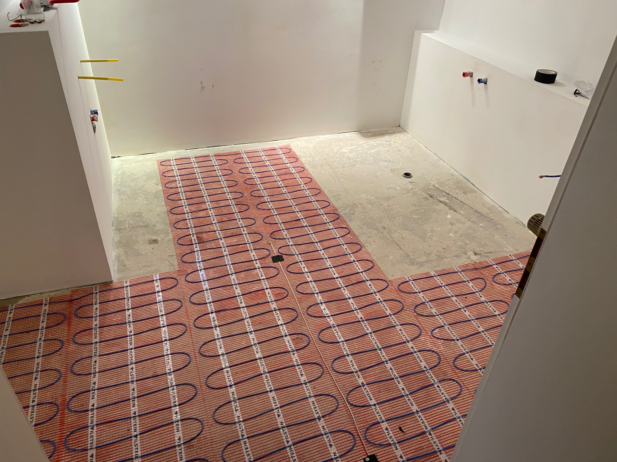 Rozłożona mata grzewcza z niebieskimi przewodami i czerwonymi elementami grzejnymi na betonowej posadzce w łazience przed położeniem płytek, widoczne rury i instalacje na ścianach.