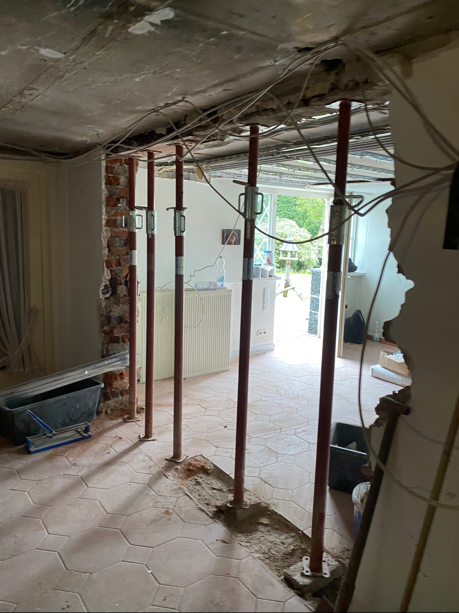 Wnętrze w trakcie przebudowy: widoczne podpory stropowe, fragmenty usuniętej ściany z cegły, odsłonięte przewody elektryczne i fragmenty starej instalacji, oraz narzędzia w kuwecie na podłodze...