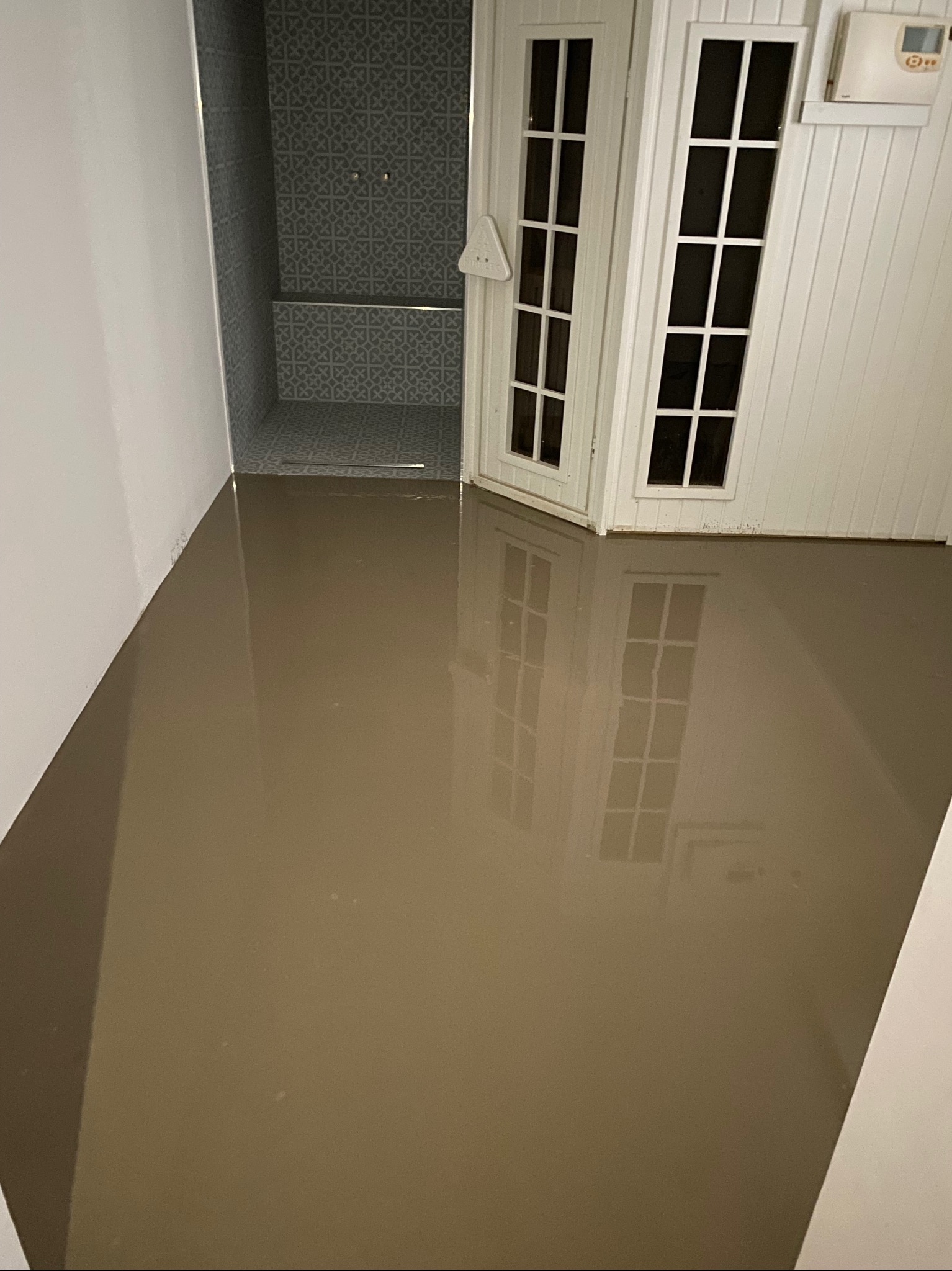 Zalana łazienka z widoczną kabiną prysznicową wyłożoną szarymi płytkami wzorzystymi i białymi drzwiami z szybkami, odbijającymi się w wodzie na podłodze.