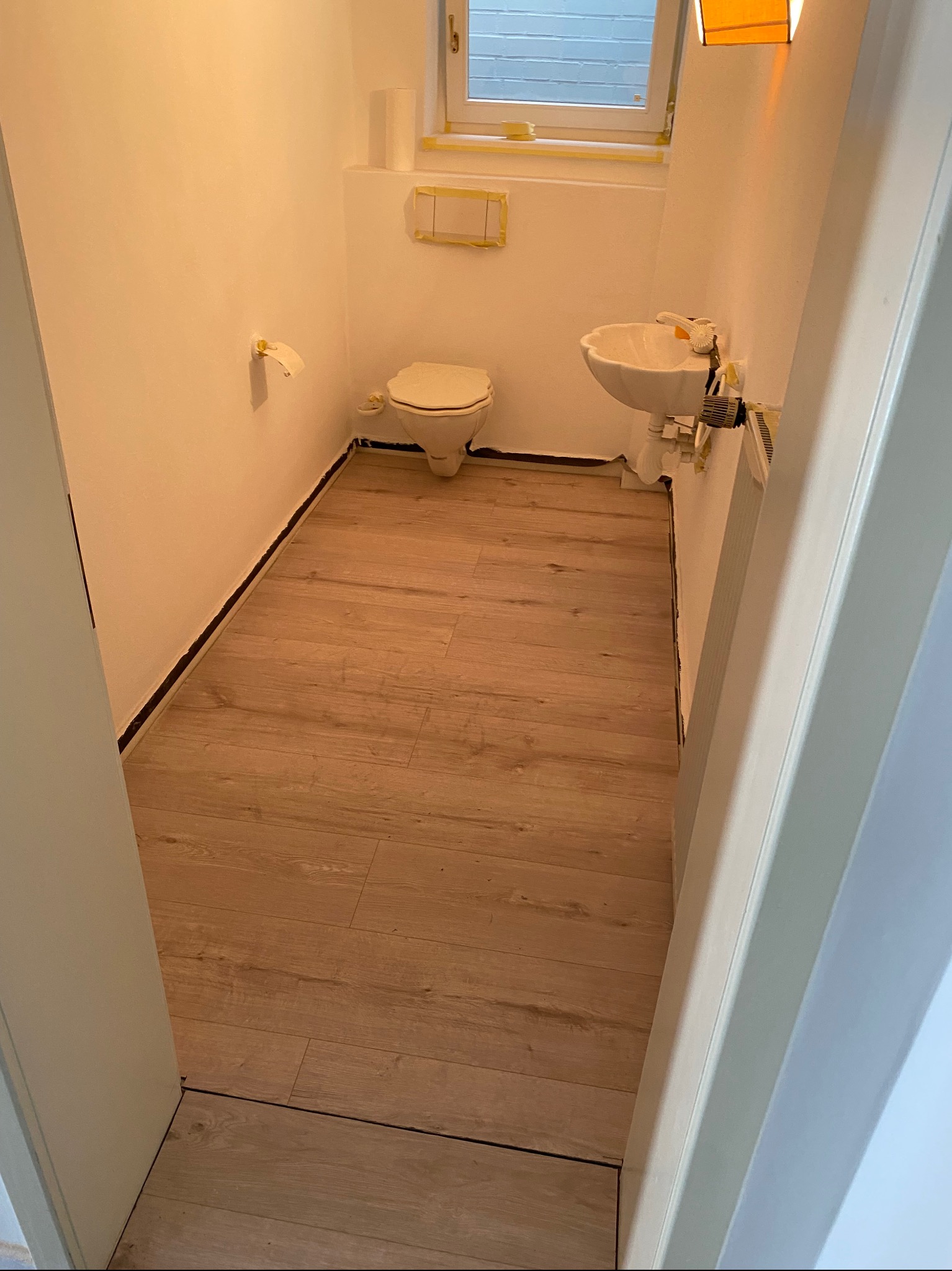 Wnętrze małej łazienki w trakcie remontu, z drewnopodobną podłogą, białymi ścianami zabezpieczonymi taśmą malarską wokół okna i toalety, oraz białą umywalką. Widoczne ślady po poprzednich pracach...