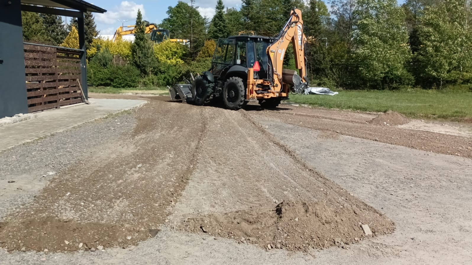 Pomarańczowo-czarna koparko-ładowarka CASE wyrównuje teren żwirowy przed posesją, widoczne ślady opon i usypane pryzmy ziemi.