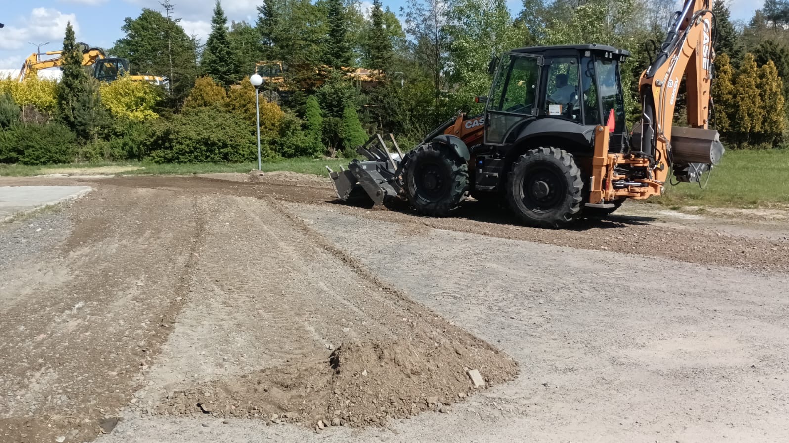 Pomarańczowo-czarna koparko-ładowarka CASE podczas wyrównywania terenu podłoża z gruzem i piaskiem, widoczne ślady opon i inne maszyny w tle.
