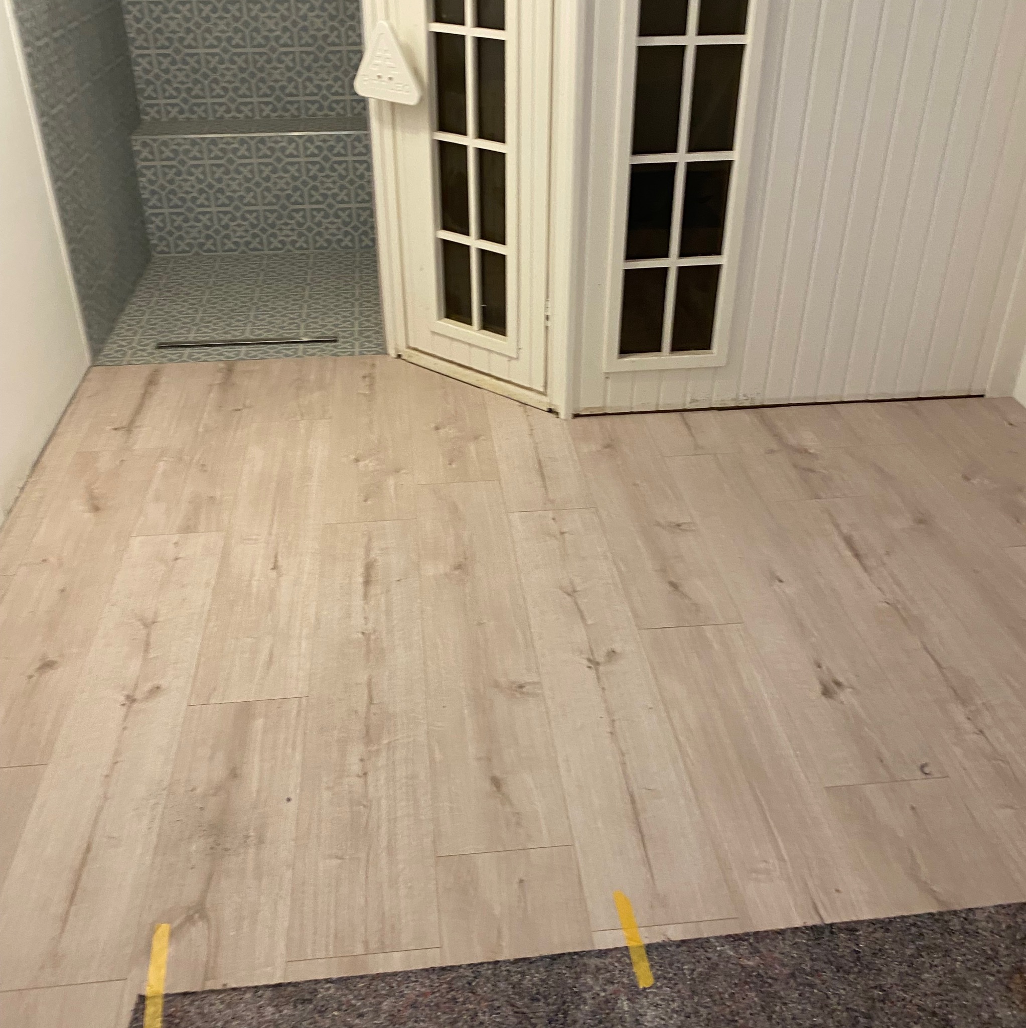 Jasne panele podłogowe ułożone w pomieszczeniu z fragmentem kabiny prysznicowej w tle i białymi panelami ściennymi oraz białymi drzwiami.