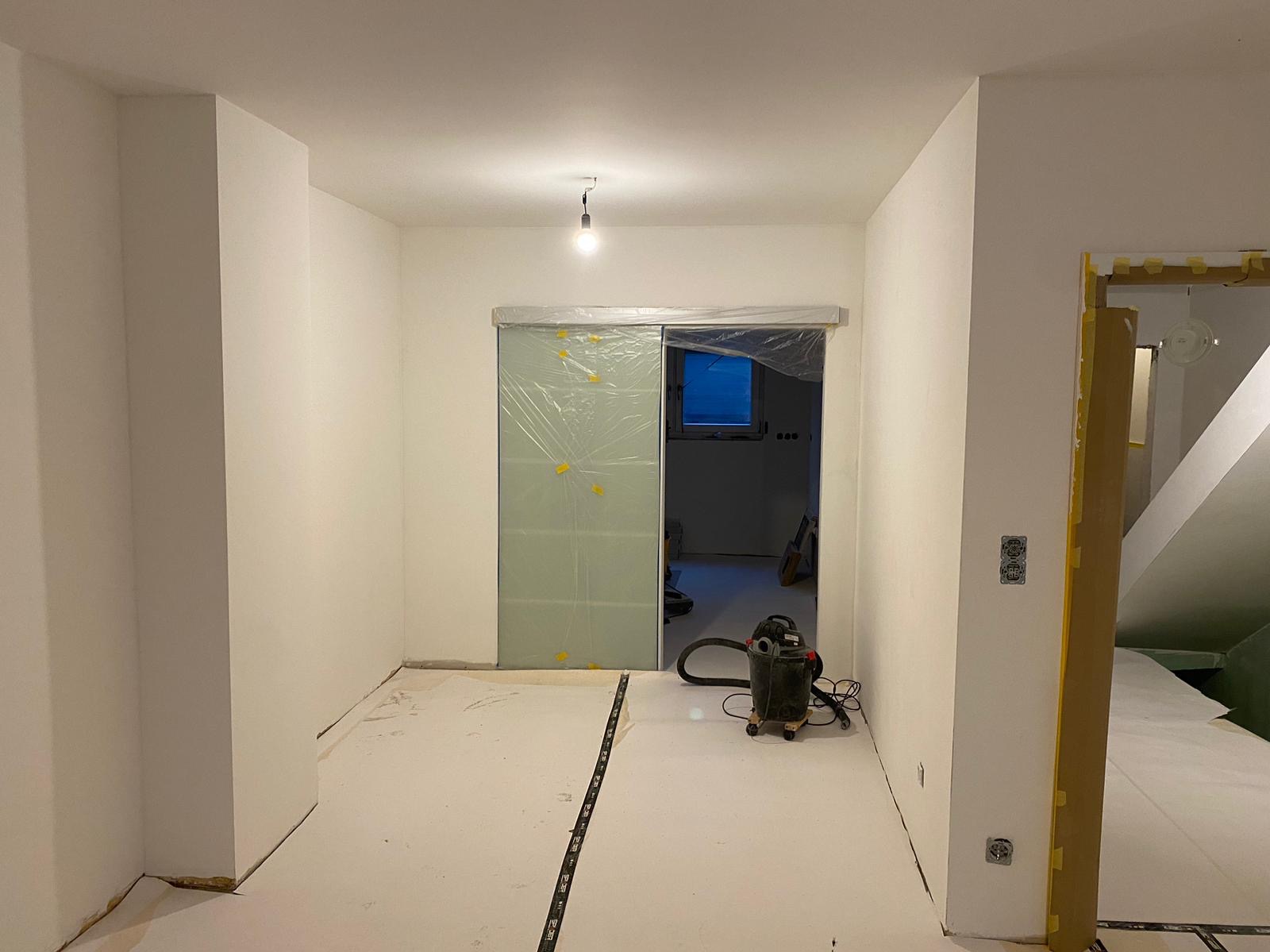 Przygotowane do malowania, białe wnętrze z zabezpieczonymi folią drzwiami przesuwnymi, podłogą i framugami, widoczny odkurzacz przemysłowy i listwa pomiarowa.
