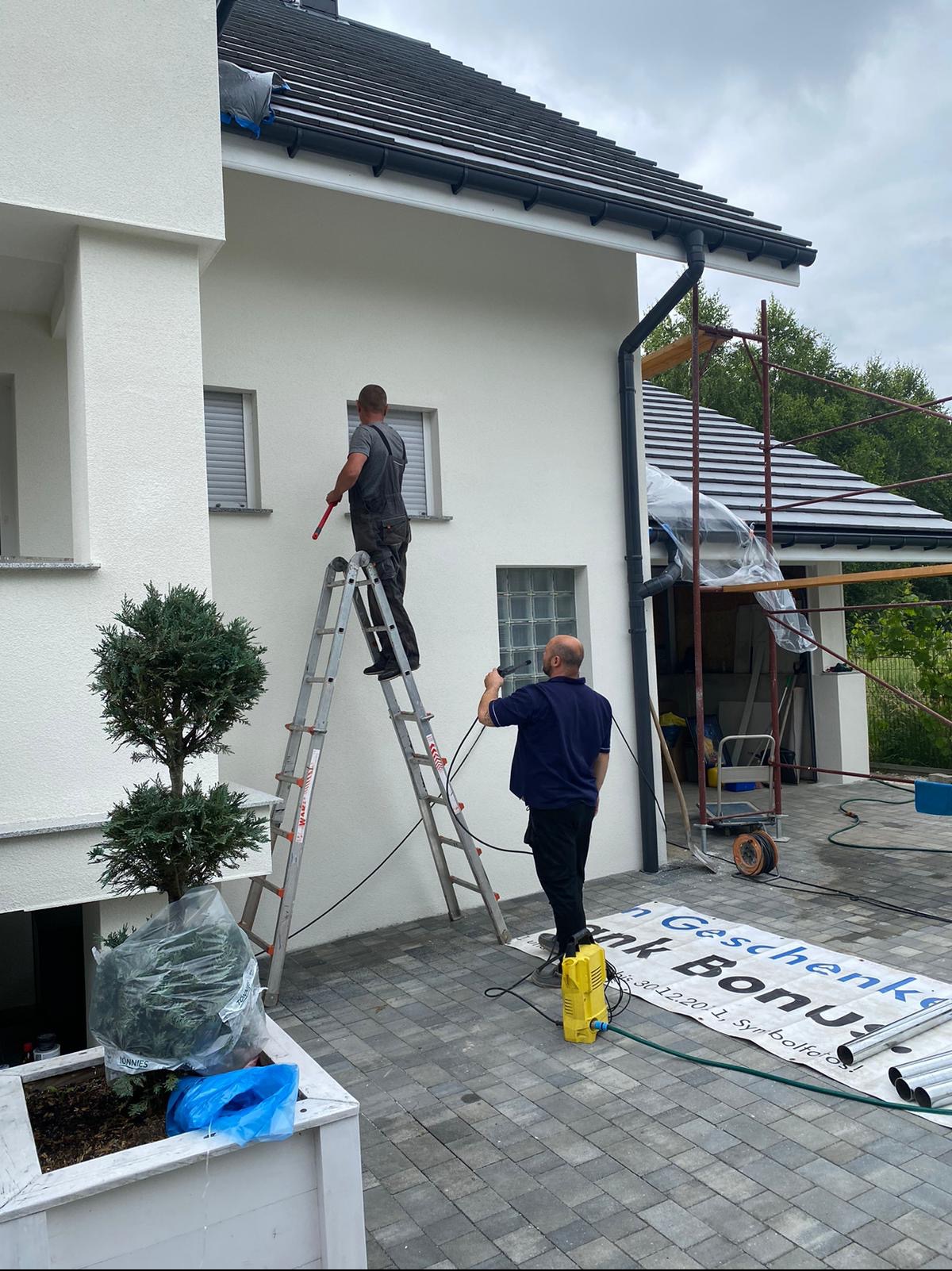Dwóch mężczyzn czyści białą elewację domu. Jeden stoi na drabinie z przedłużką do wałka, drugi obsługuje myjkę ciśnieniową na ziemi. Widoczne rusztowanie z boku budynku i donica z drzewkiem.